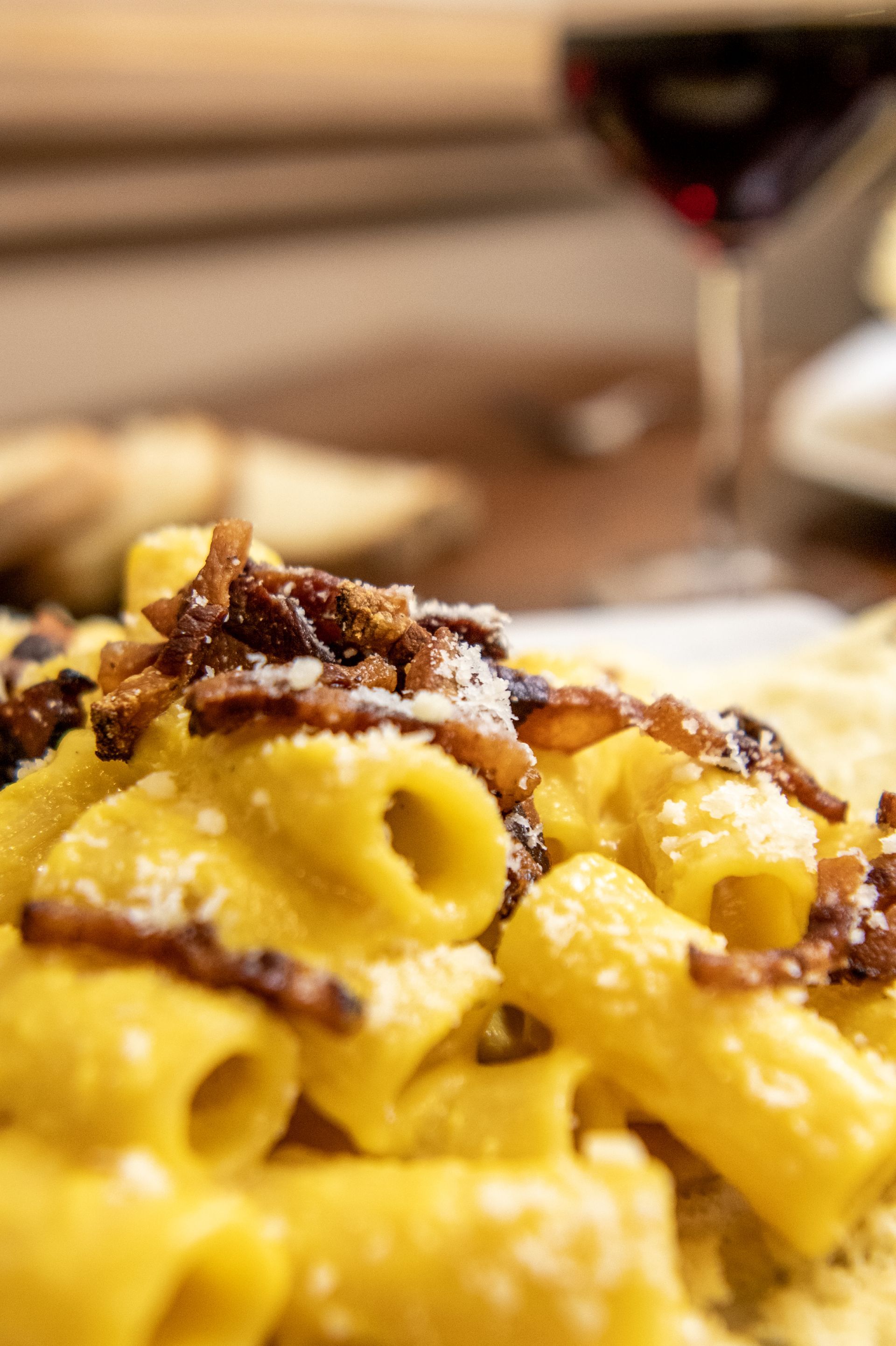Pasta con sugo e formaggio grattugiato in un piatto, con un bicchiere di vino sfocato sullo sfondo.