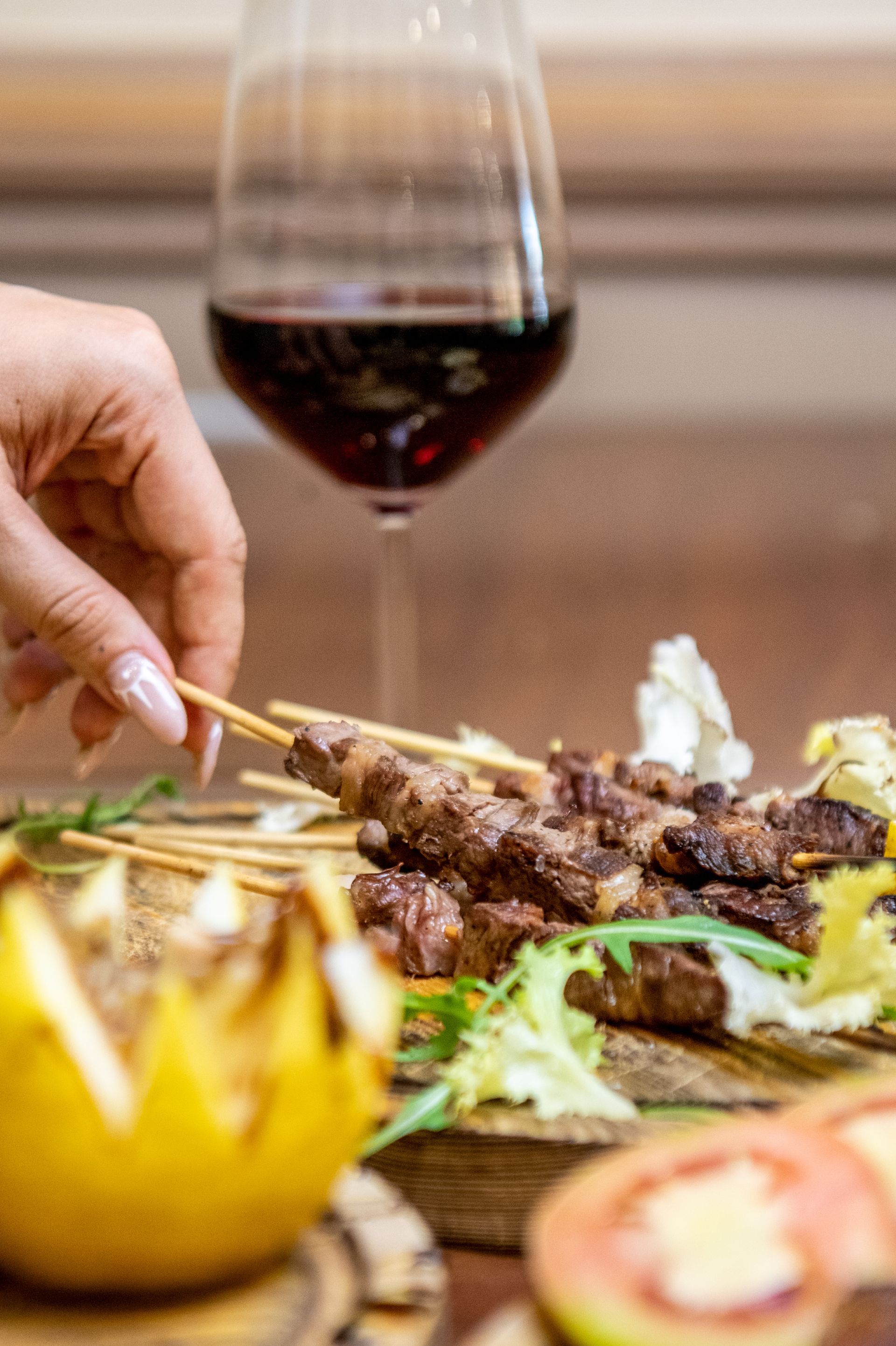 Una mano si allunga verso della carne grigliata su un piatto, con del vino rosso sullo sfondo.