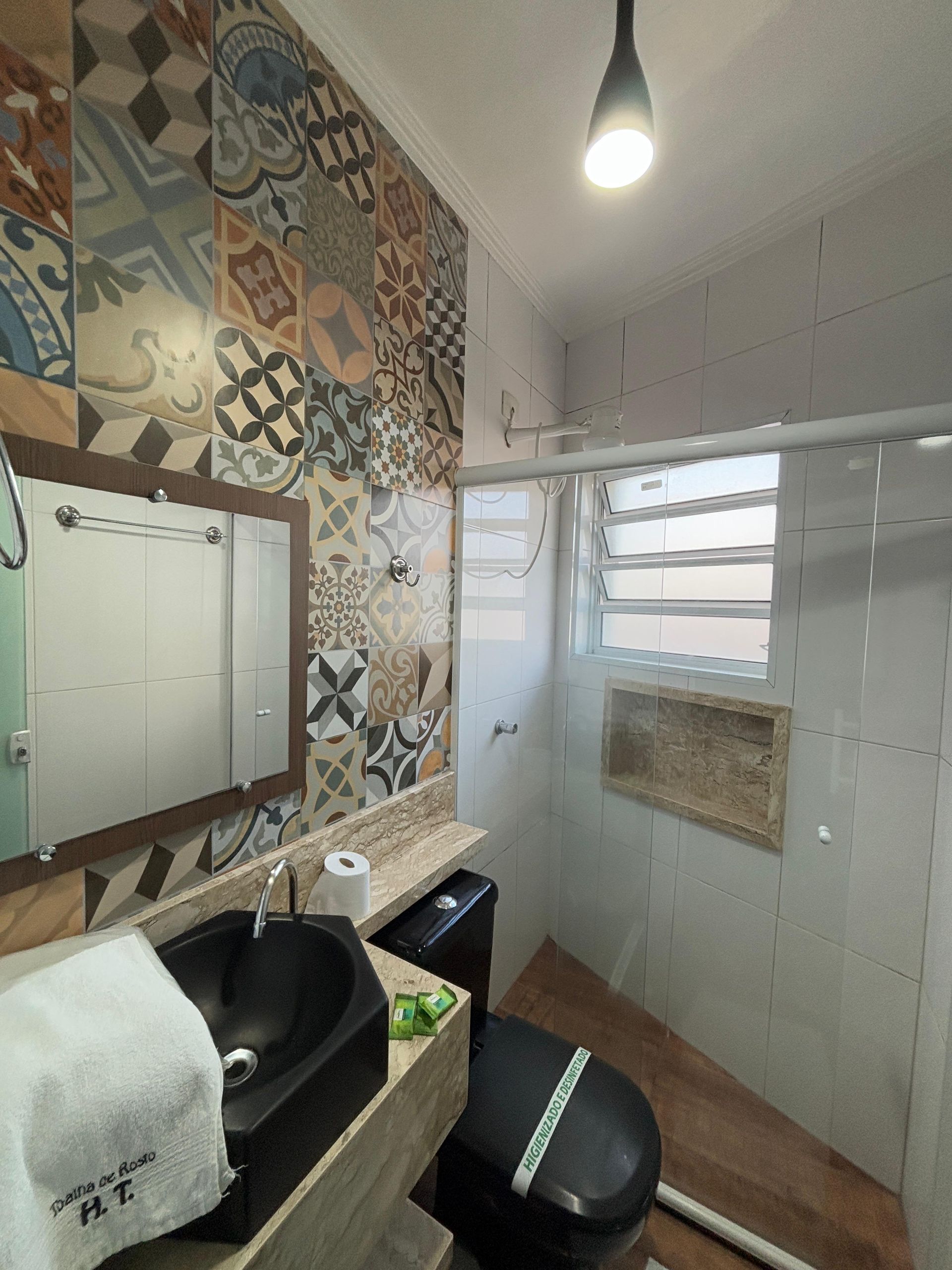 Um banheiro moderno com azulejos estampados nas paredes, uma pia de apoio preta, um vaso sanitário, uma bancada de madeira e um box de vidro para o chuveiro.