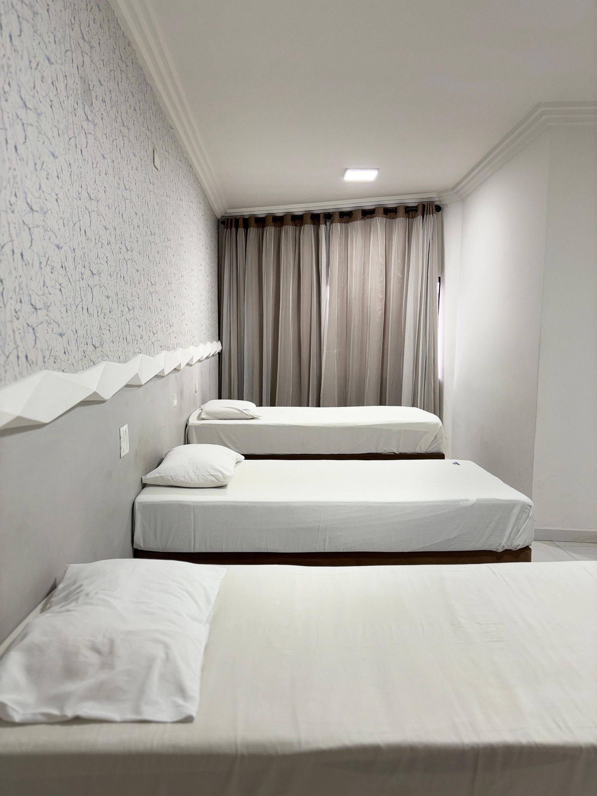Um quarto de hotel com três camas brancas enfileiradas, papel de parede cinza estampado e cortinas cinza que vão até o chão.