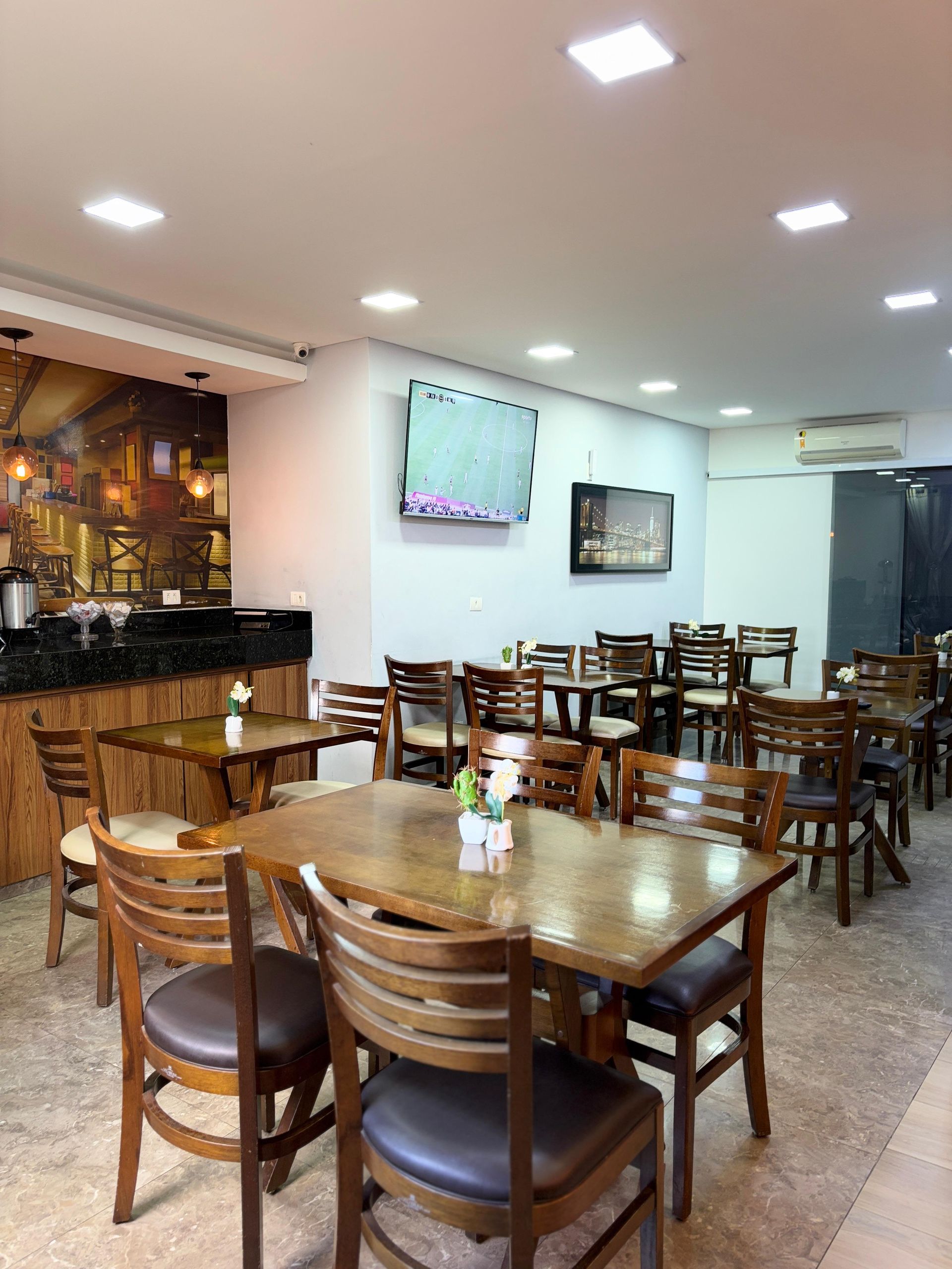Interior de restaurante com várias mesas e cadeiras de madeira dispostas sobre um piso salpicado sob iluminação embutida.