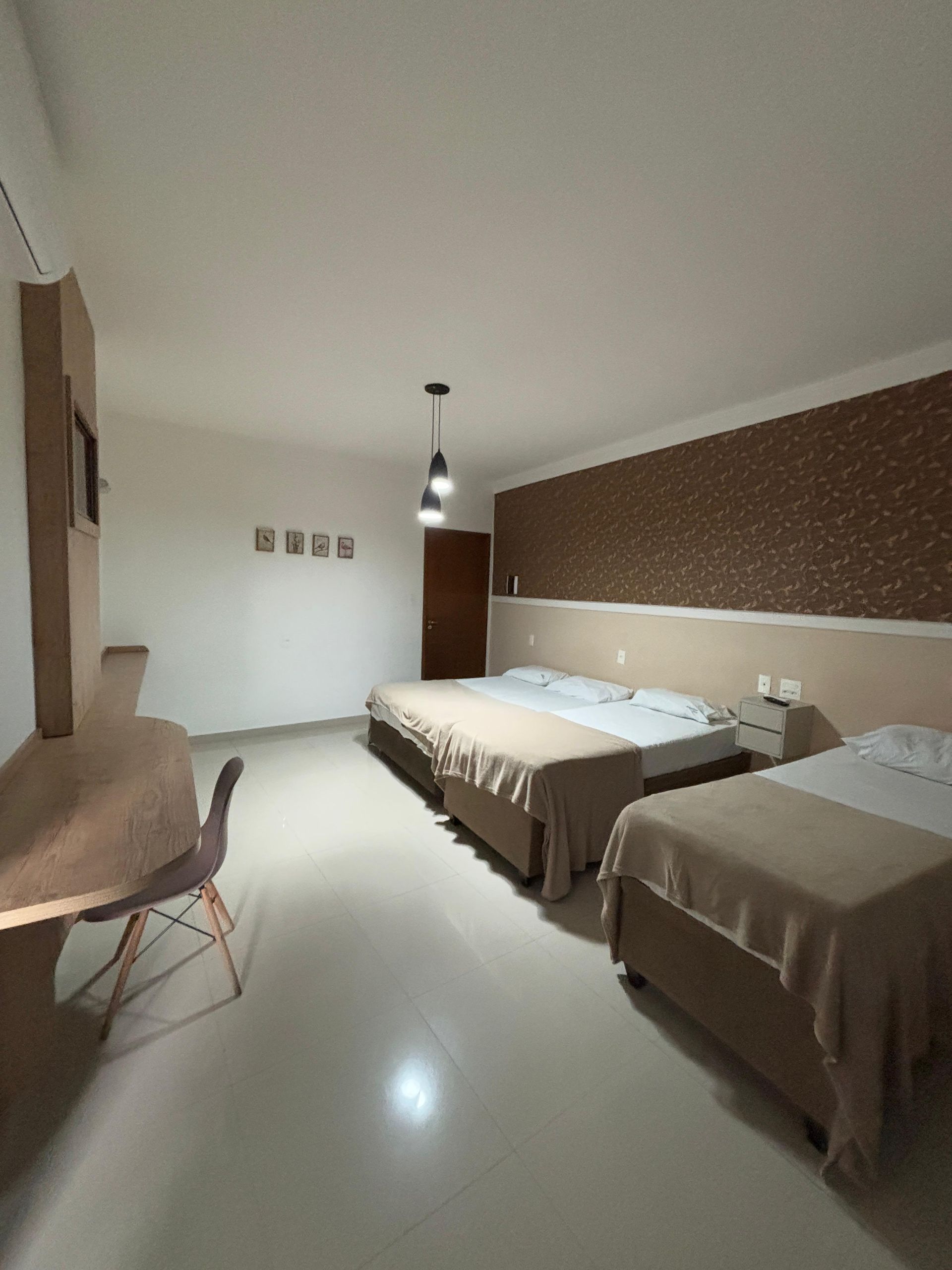 Quarto de hotel com três camas, escrivaninha e uma parede com detalhes em tijolo marrom.