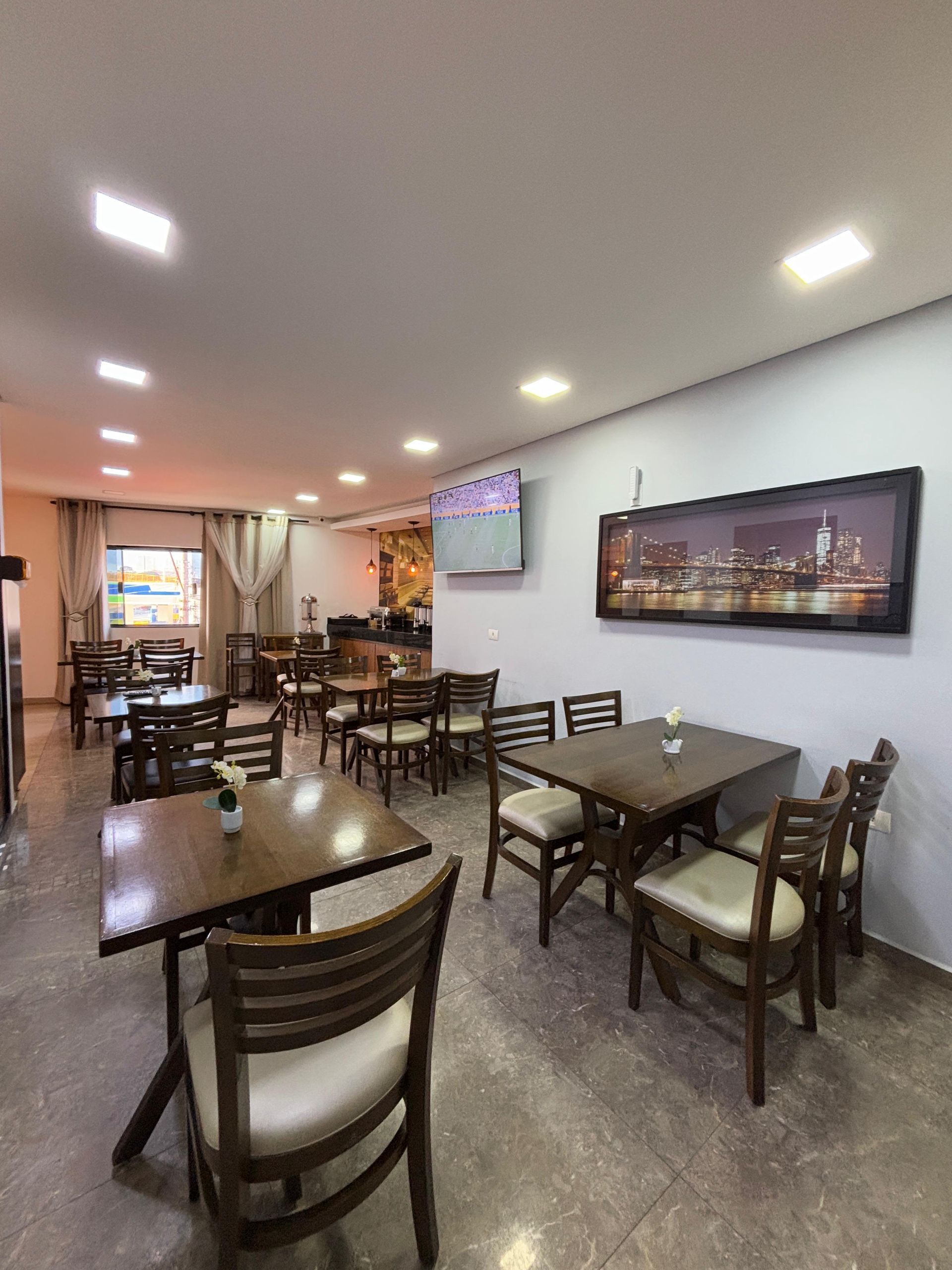 Interior de restaurante com mesas e cadeiras de madeira, uma pintura de paisagem urbana e uma pequena TV.