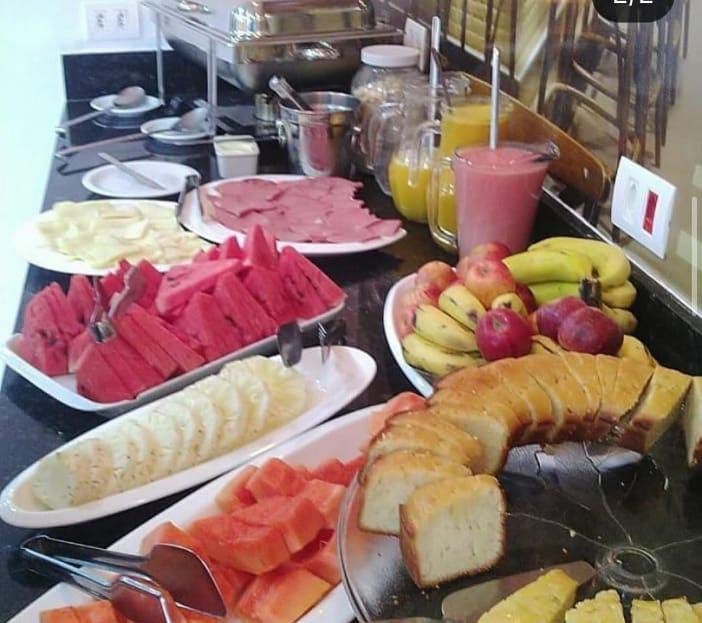 Buffet de café da manhã com frutas fatiadas, pão, suco e frios em uma bancada preta.
