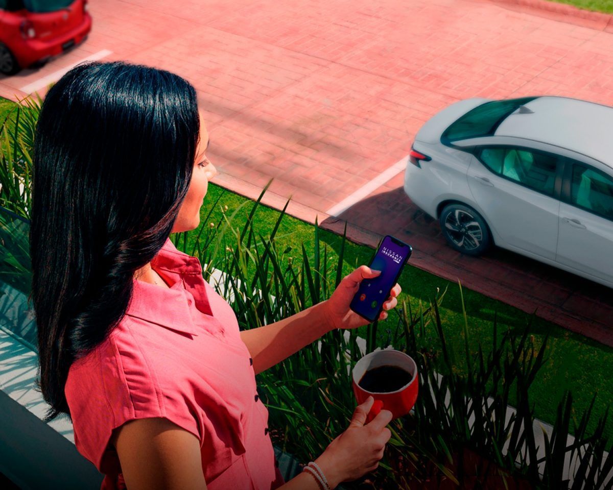 Una persona con una camisa rosa sostiene un teléfono y una taza, mirando un coche blanco que pasa desde un balcón.