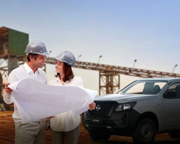 Dos ingenieros con cascos revisan planos junto a una camioneta negra en una obra de construcción.