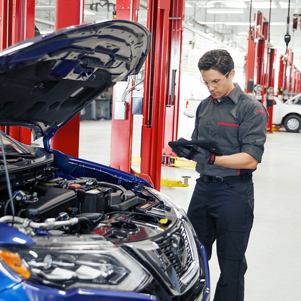 Un mecánico en un taller rojo inspecciona un coche azul con el capó abierto usando una tableta.
