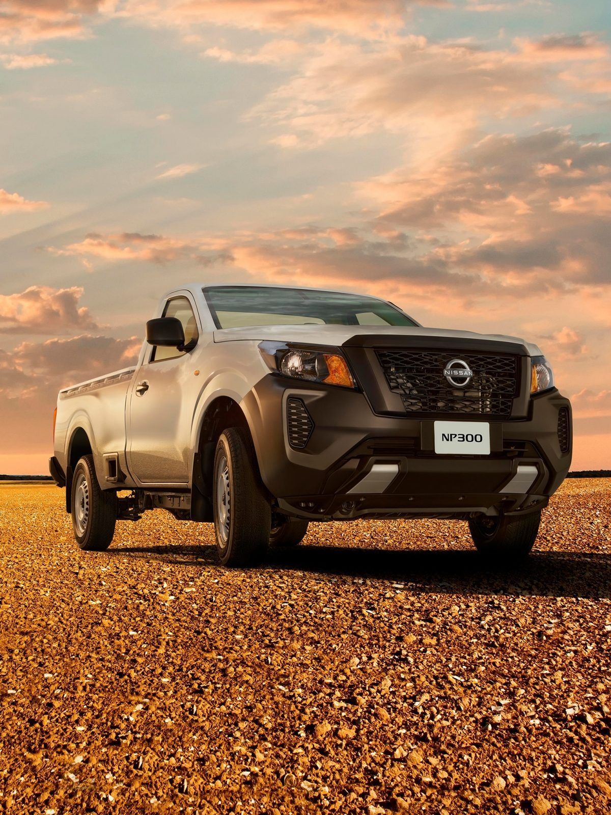 Camioneta Nissan blanca estacionada en una playa de grava al atardecer bajo un cielo nublado de color naranja.