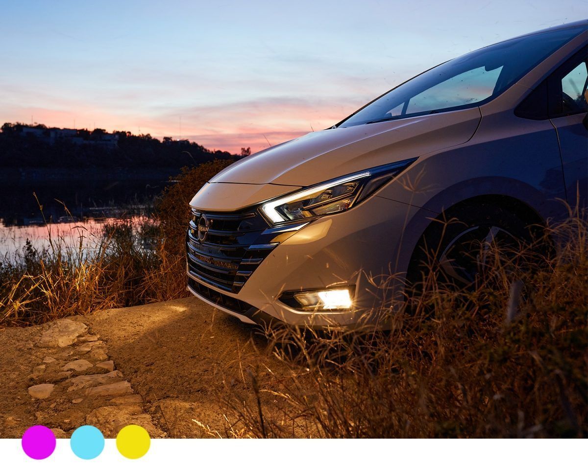 Coche plateado aparcado junto a un río al atardecer, con cálidos reflejos del cielo y hierba seca en primer plano.