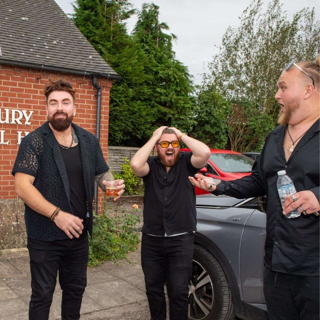 Three people stand outdoors near a brick building; one has hands on his head, another holds a drink, and one holds a water.