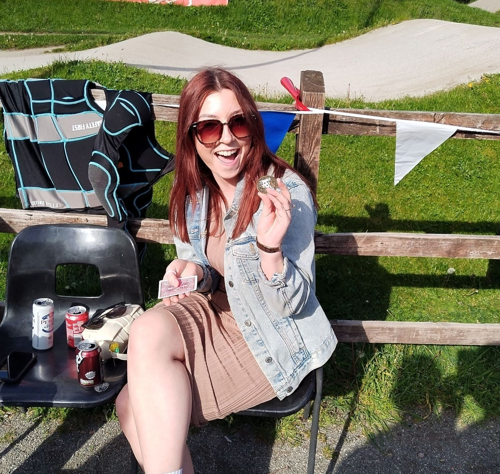 Woman in sunglasses and denim jacket, seated and smiling, holds an object in her hand