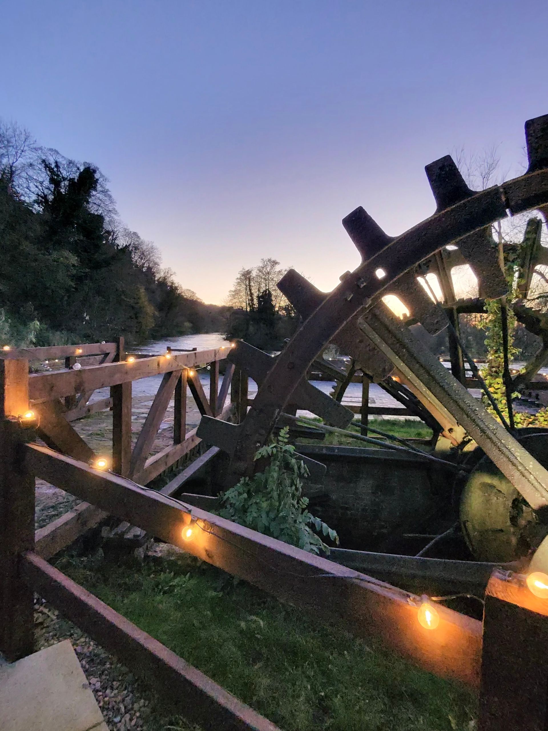 Wooden water wheel and fence with lights, set against a river at dusk.