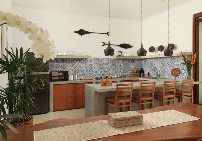 Kitchen with blue patterned backsplash, concrete counters, wooden cabinets and chairs, and decorative lighting.