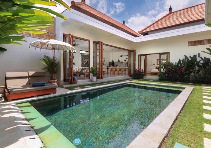 Pool in a courtyard, flanked by a lounge chair with an umbrella, and surrounded by a house with open doors.