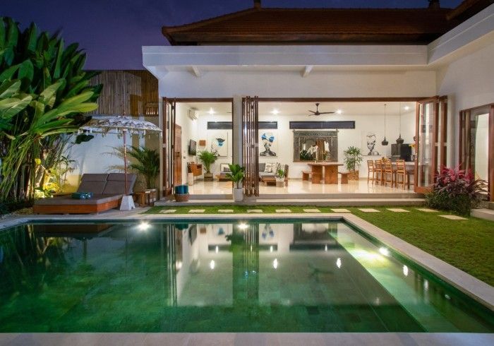 Poolside villa at dusk, with open doors to the lit living space and a green-tinted pool.