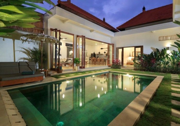 Pool and villa exterior at dusk; green pool, white walls, open doors, red roof, lounge area.