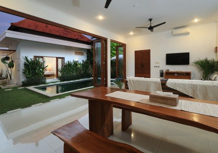 Open-plan interior with wood dining table, pool view. White walls, wood trim, lush greenery.