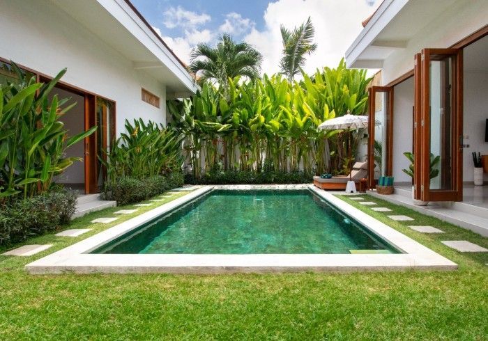 A rectangular swimming pool surrounded by lush greenery, framed by a white-walled villa with open wooden doors.