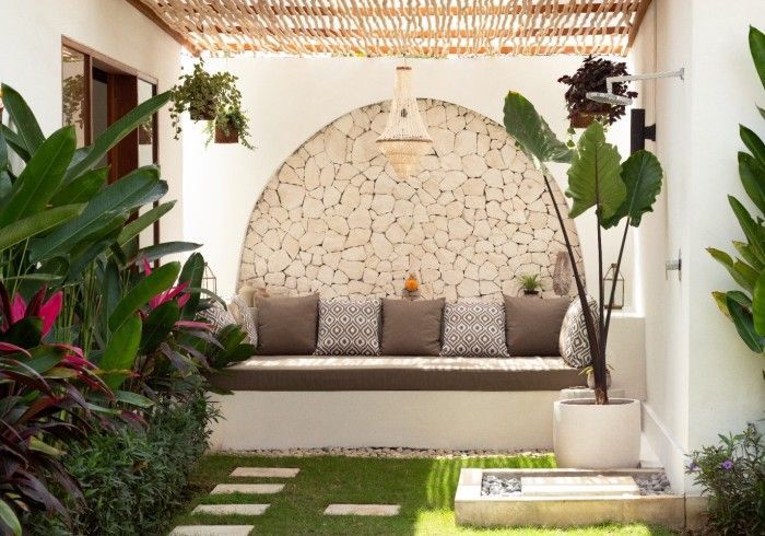Outdoor lounge area with stone wall, built-in bench with pillows, plants, and a woven ceiling.