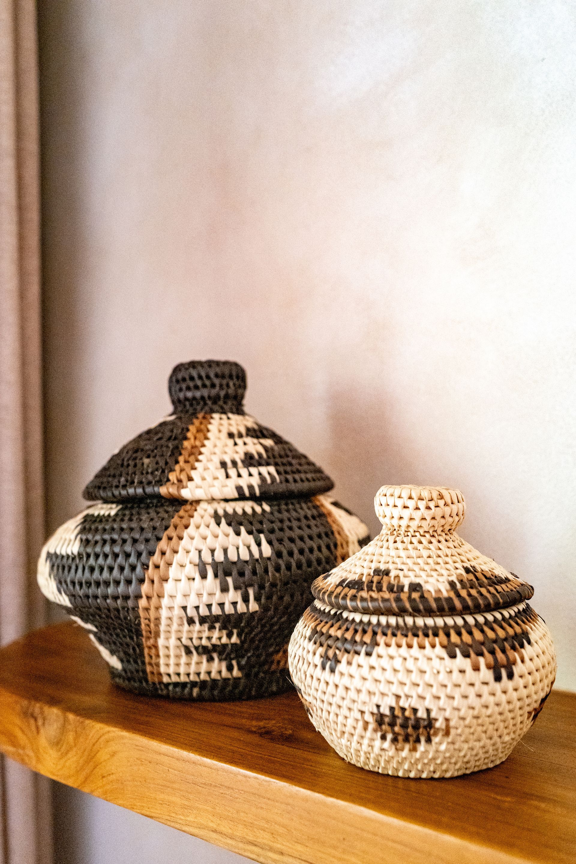 Two woven lidded baskets on a wooden shelf, beige and dark patterned.