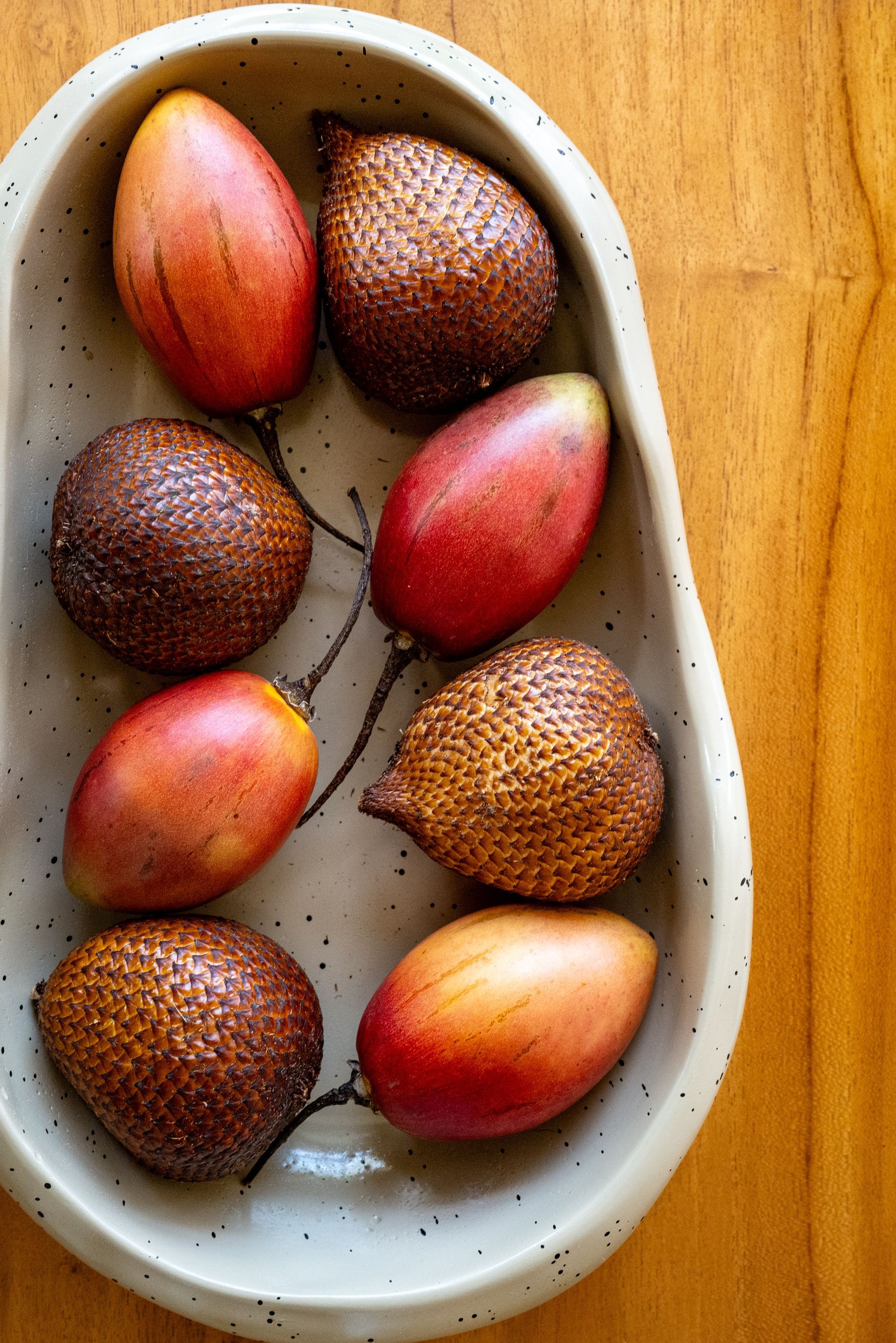 Oval dish with red tamarillos and brown snake fruit on a wooden table.