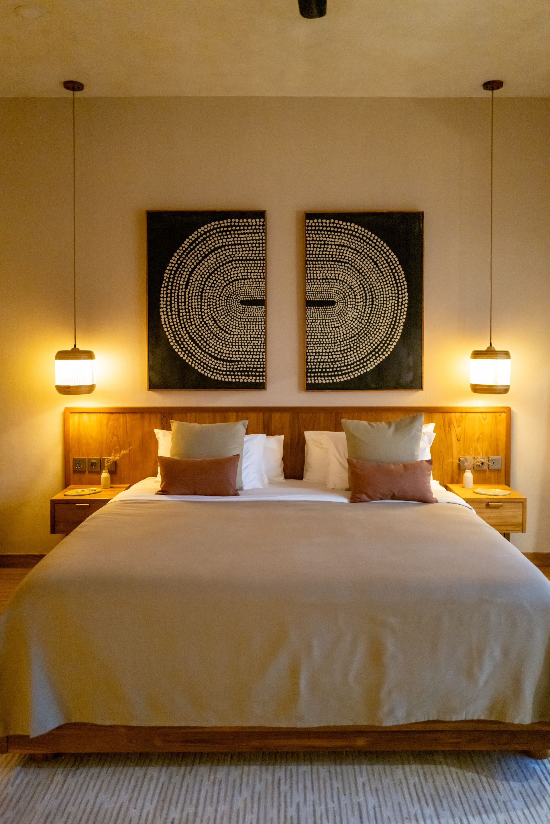 Bedroom with a bed, two abstract artworks, and hanging lamps. Earthy tones with wooden accents.
