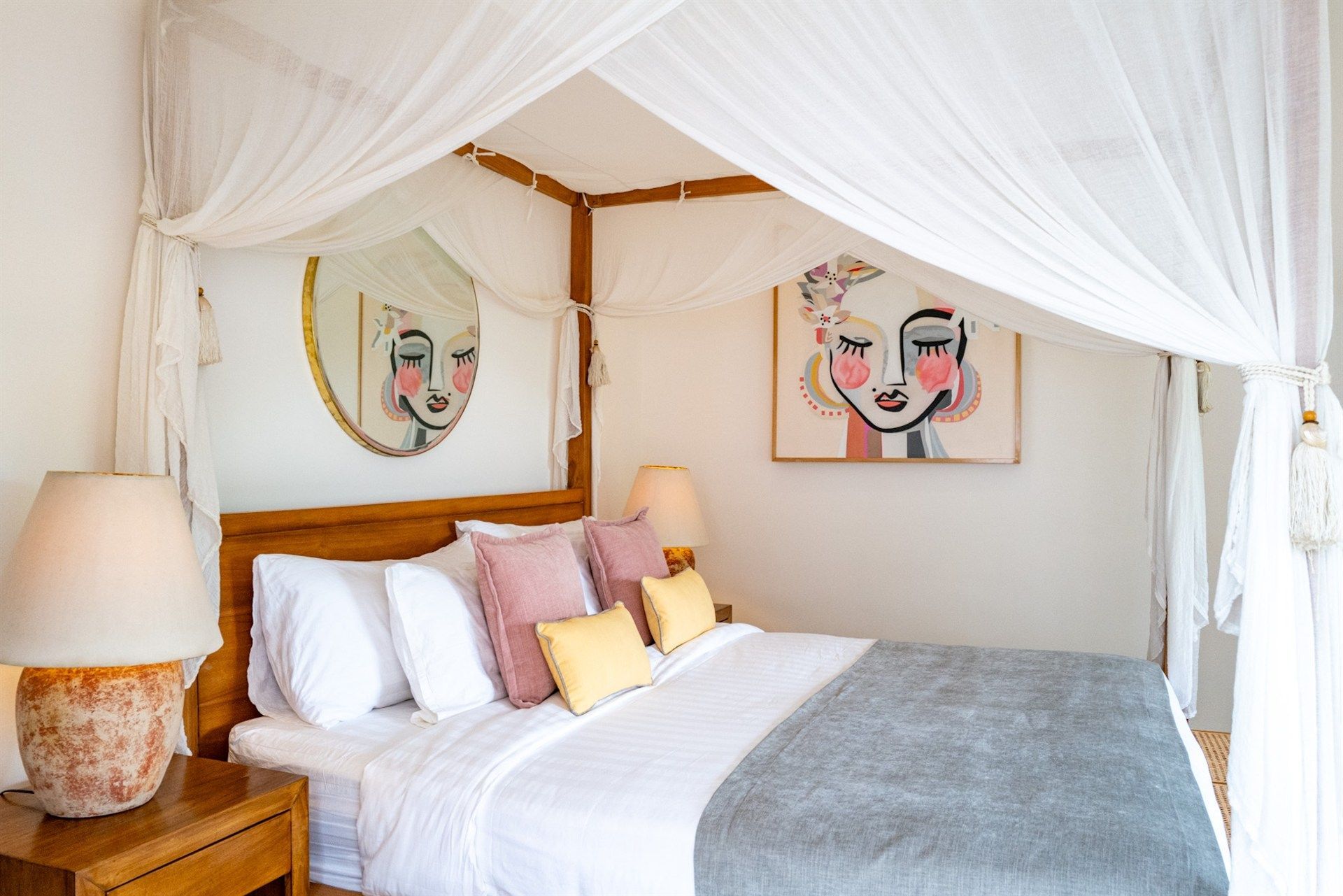Bedroom with four-poster bed, white canopy, decorative pillows, artwork, and a bedside lamp.