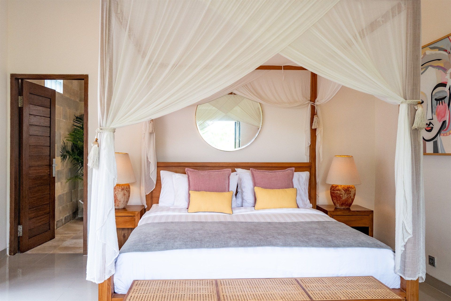 Bedroom with four-poster bed, white canopy, bedside tables, mirror, and artwork on the wall. Open door to bathroom.