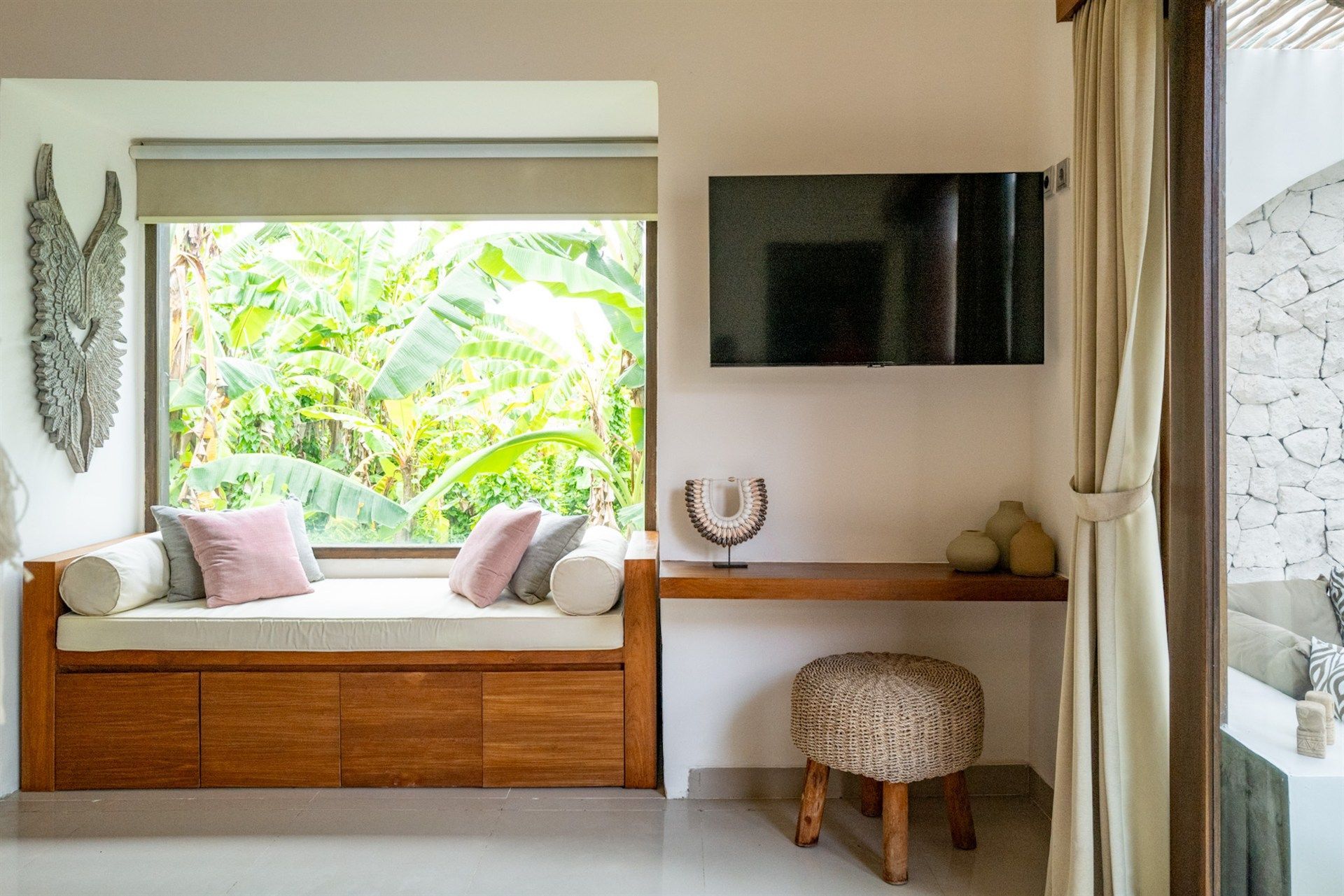 A room with a window seat, desk, and TV. The window overlooks lush greenery. A woven stool sits below the desk.