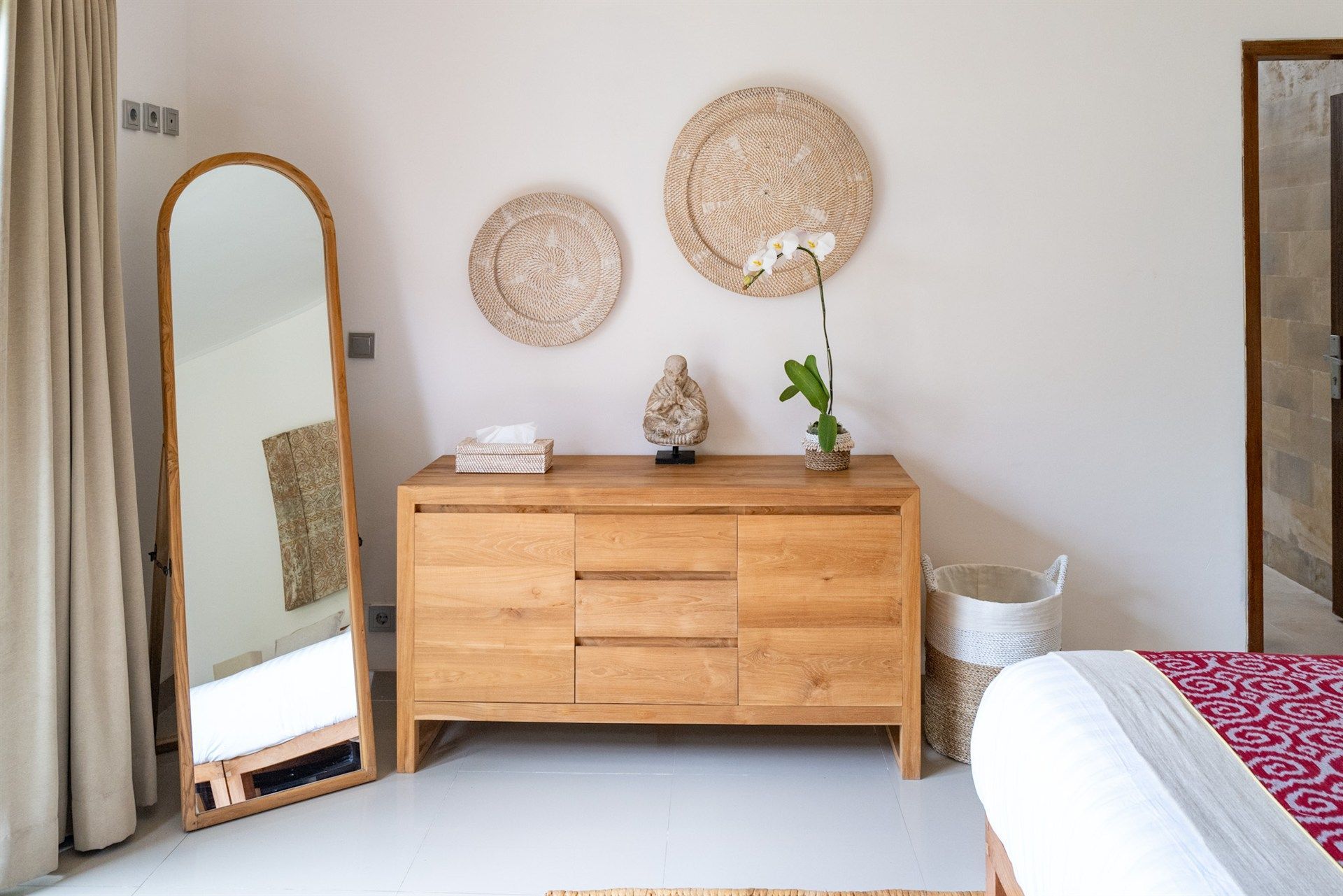 Wooden dresser with decorative woven plates, full-length mirror, vase, and bed in a bright room.