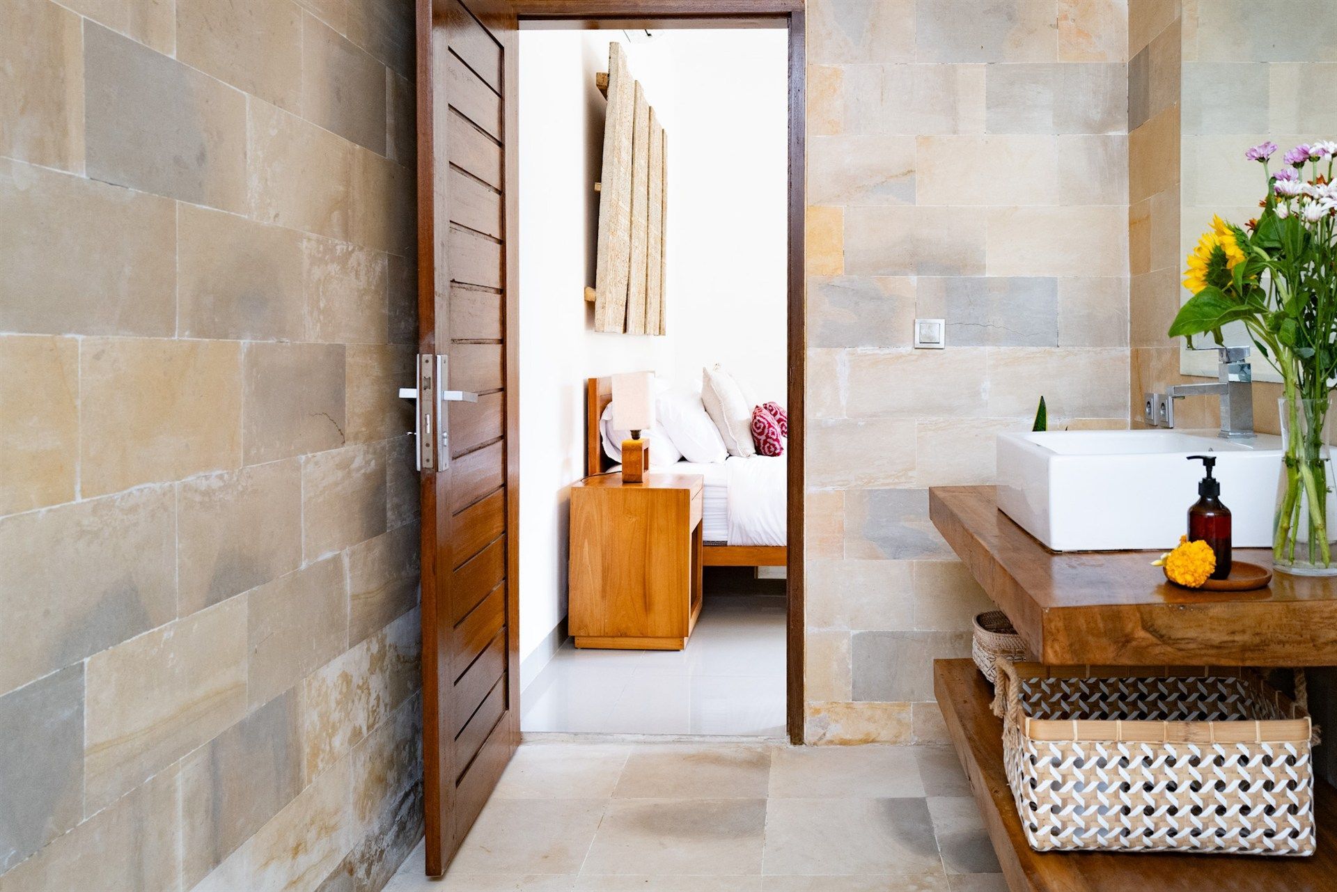 Bathroom interior with open doorway to bedroom. Stone walls, wooden shelves with sink, sunflower bouquet.