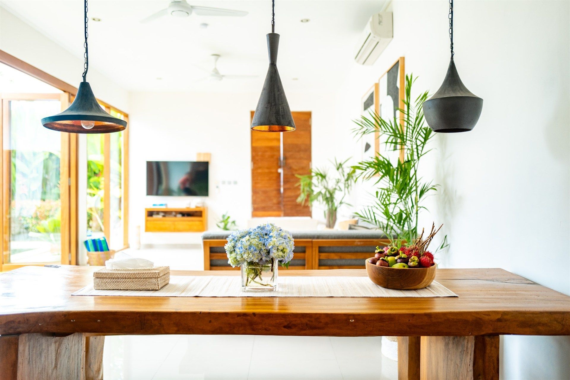 Wooden dining table with blue flowers, fruit bowl, and pendant lights. Bright room with sliding glass doors.