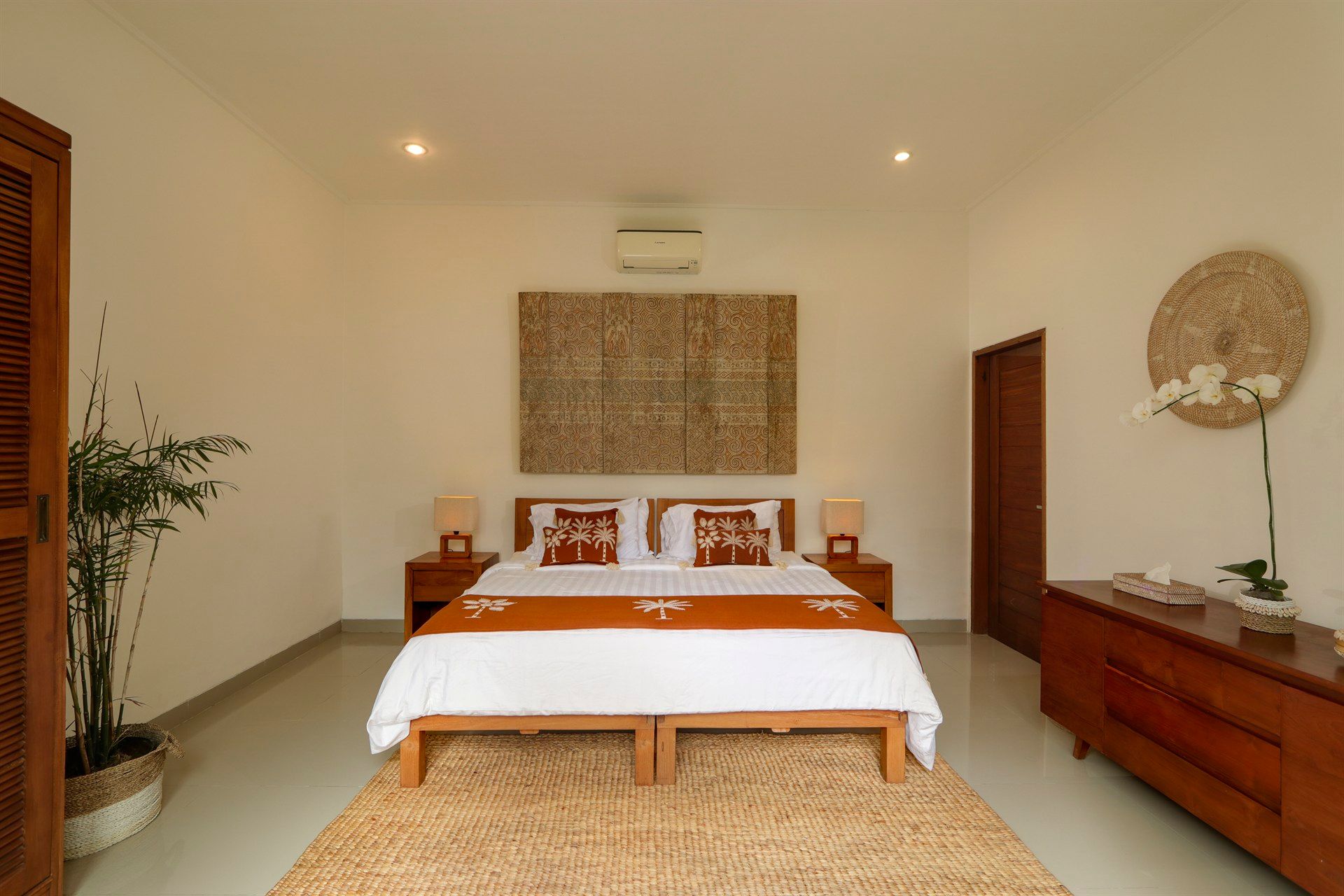 Bedroom with wooden furniture, bed, and decorative rug. White walls, warm lighting, and a woven wall hanging.