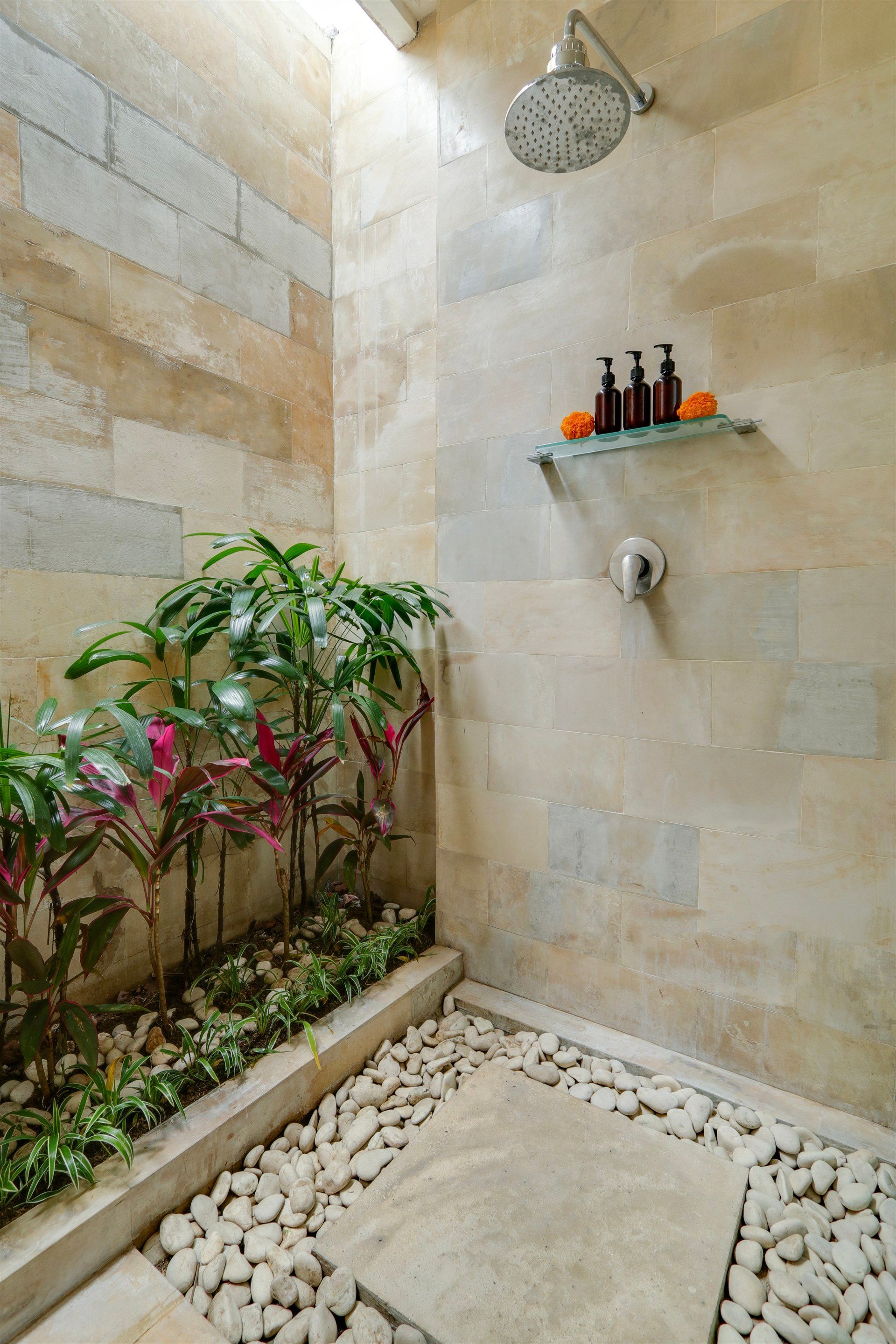 Stone-walled outdoor shower with plants in a bed of white pebbles; shampoo bottles on a glass shelf.