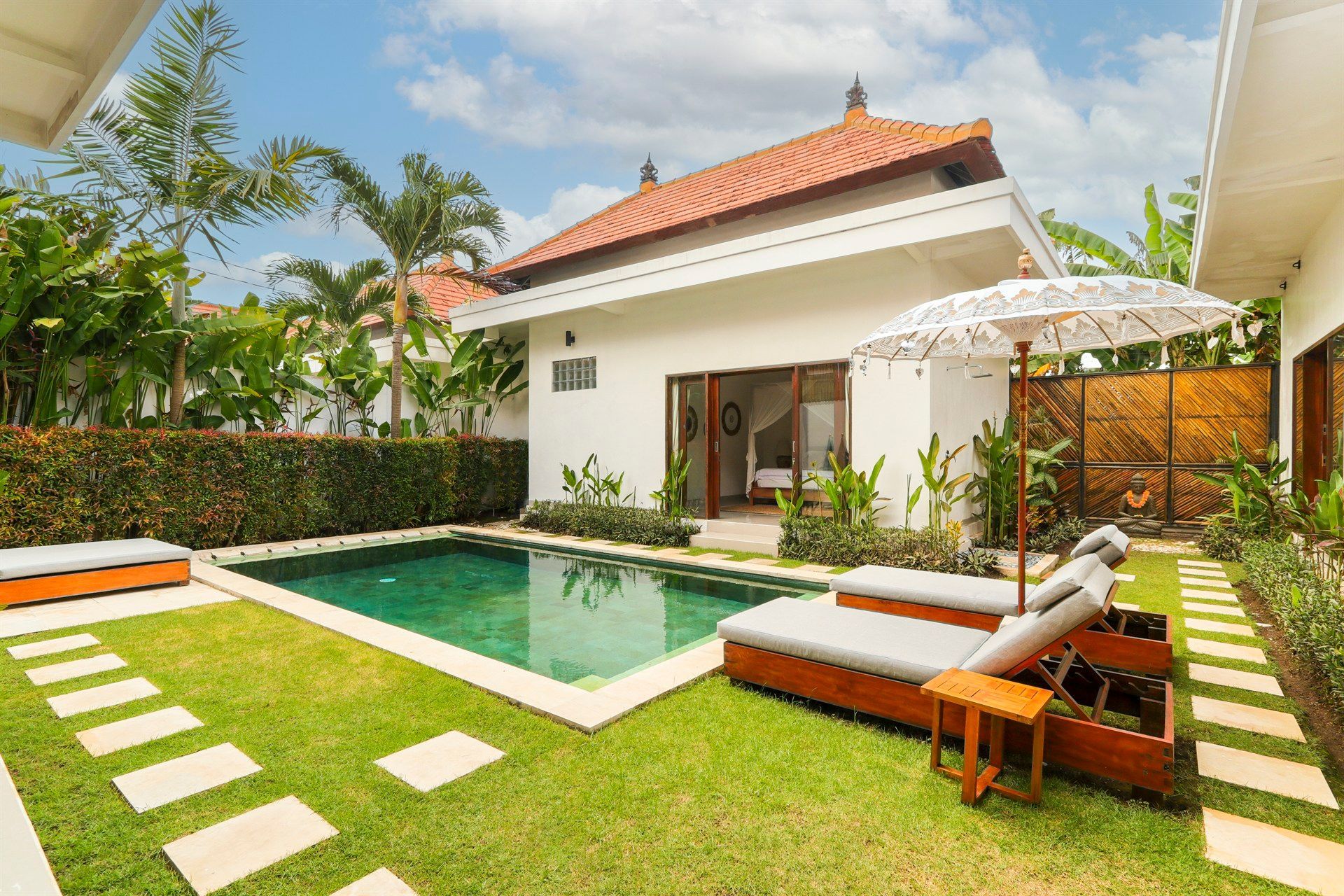 Small pool in a tropical backyard with lounge chairs, umbrella, and a white building.