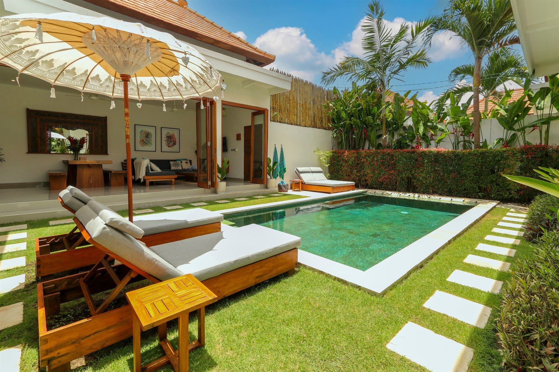 Backyard with pool, lounge chairs, umbrella, and a glimpse of an open-air living space in a tropical setting.