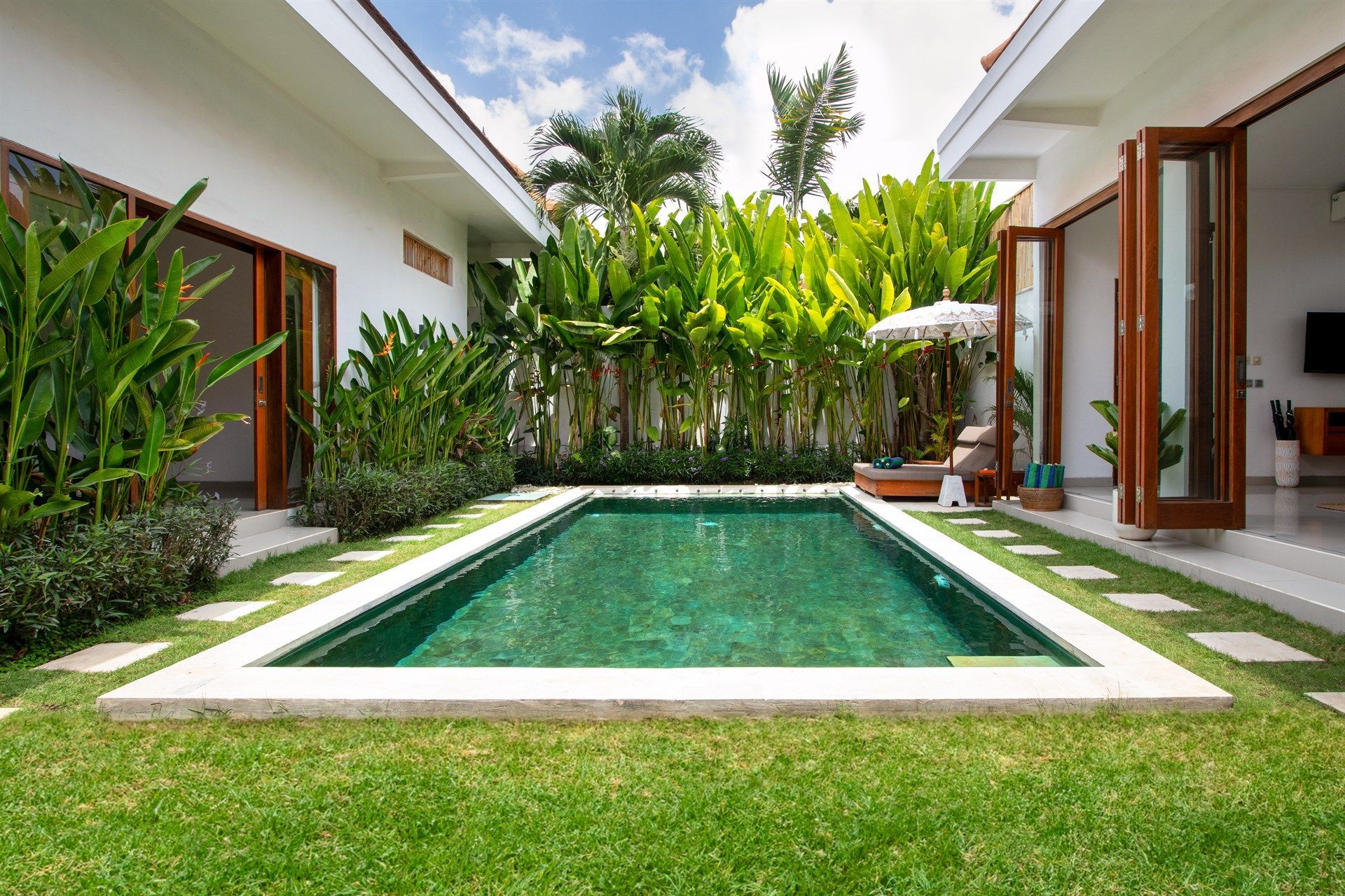 A rectangular turquoise pool in a courtyard with green grass, white walls, and tropical plants.