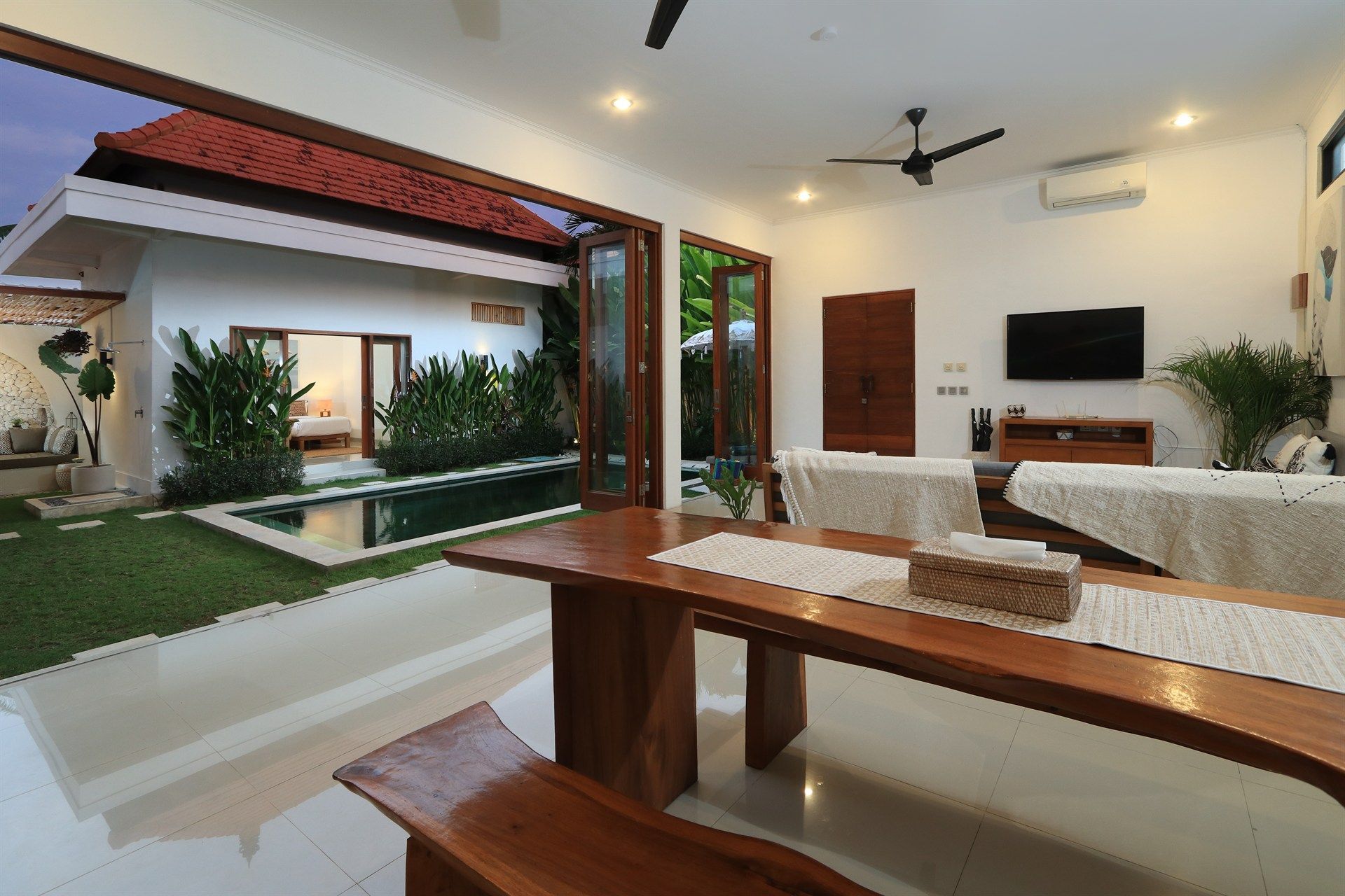 Dining room with open doors to a pool and garden; wooden table, bench, white walls, and TV.