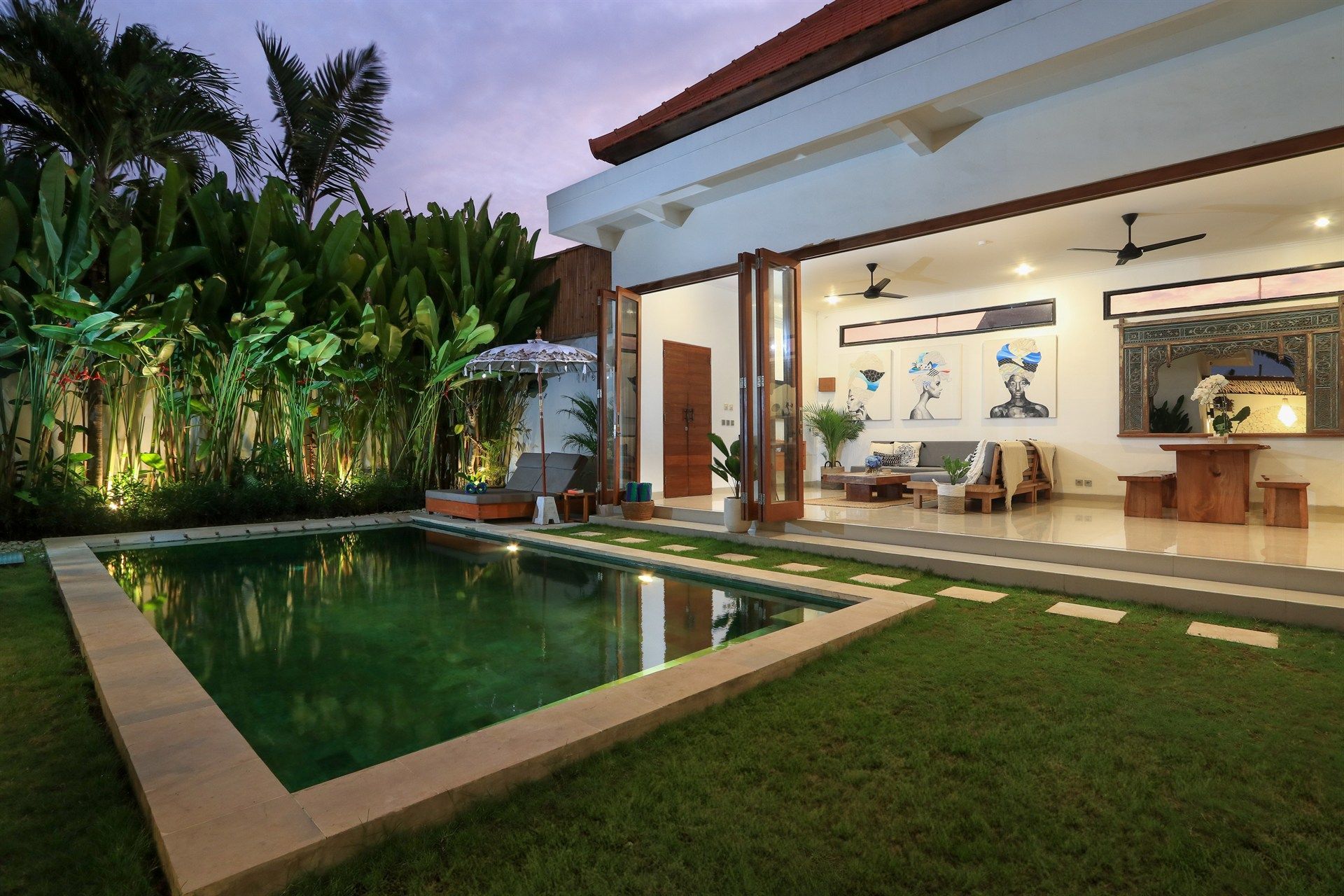 Poolside villa with a green pool, lush lawn, and open-air living space at twilight.
