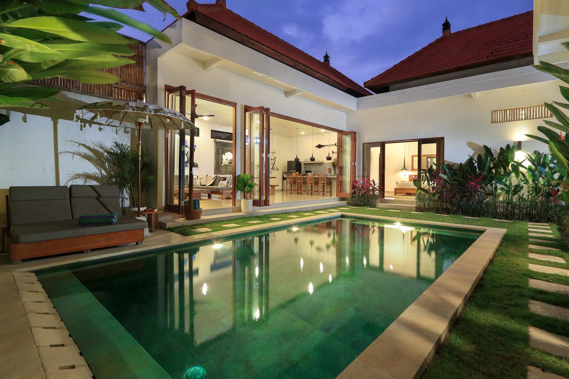 Swimming pool in front of a villa at dusk with open doors, green grass, and lounge chairs.