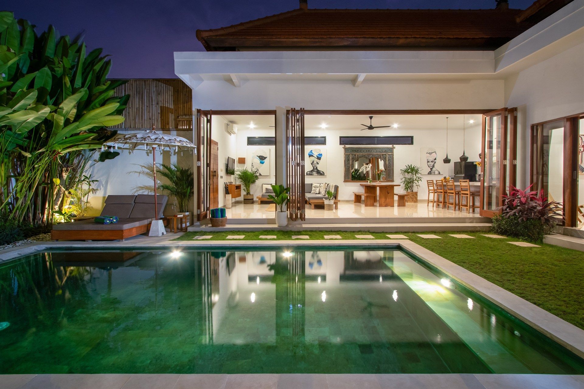 Night view of a villa with an illuminated pool and open doors to the interior, lush greenery surrounding.