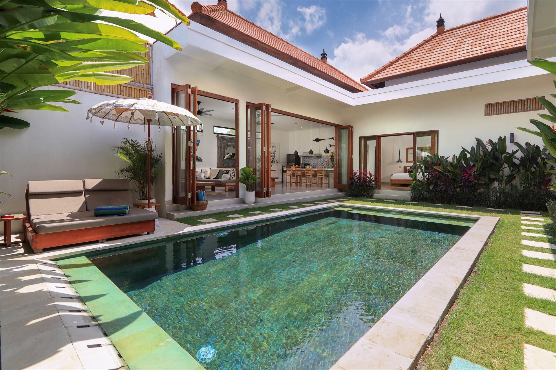 Courtyard with pool, villa, lounge chairs, and open doors to living areas, tropical plants, and bright sunshine.