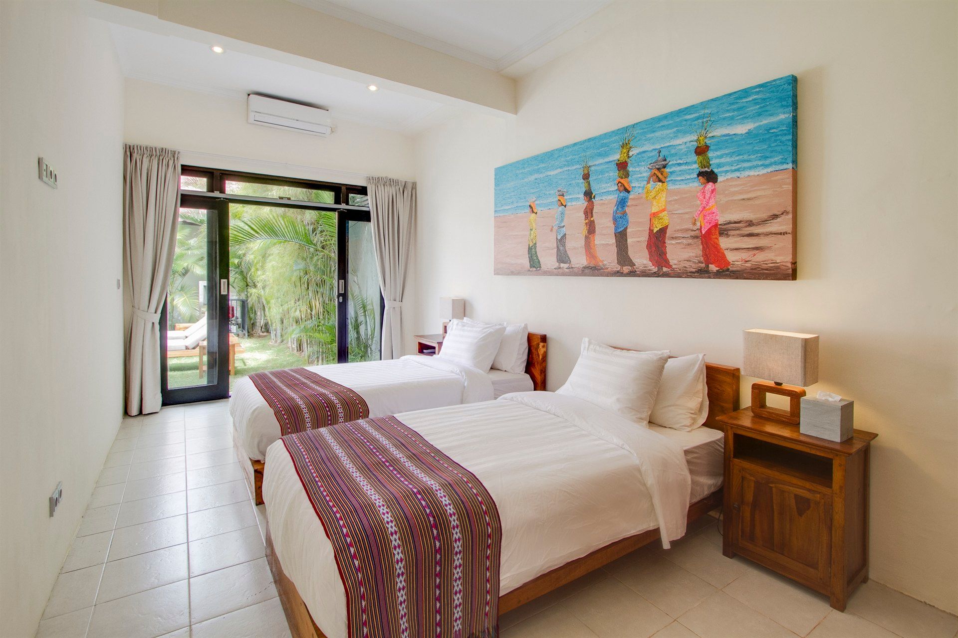 Twin beds in a brightly lit room with a colorful painting and open doors to a garden.