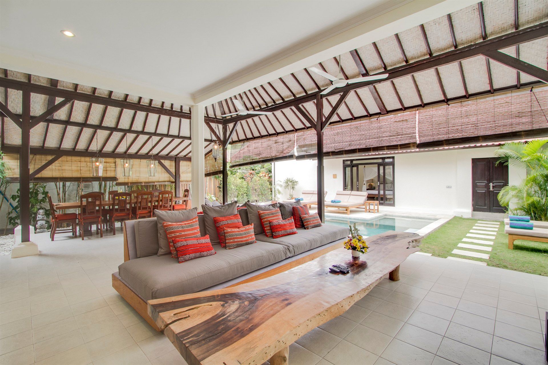 Open-air living area with a long wooden sofa and a dining table, overlooking a pool and green lawn.