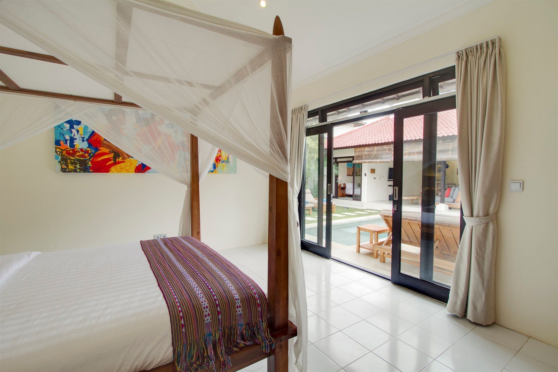 Bedroom with canopy bed, open sliding doors to a patio. White tiled floor, neutral walls.