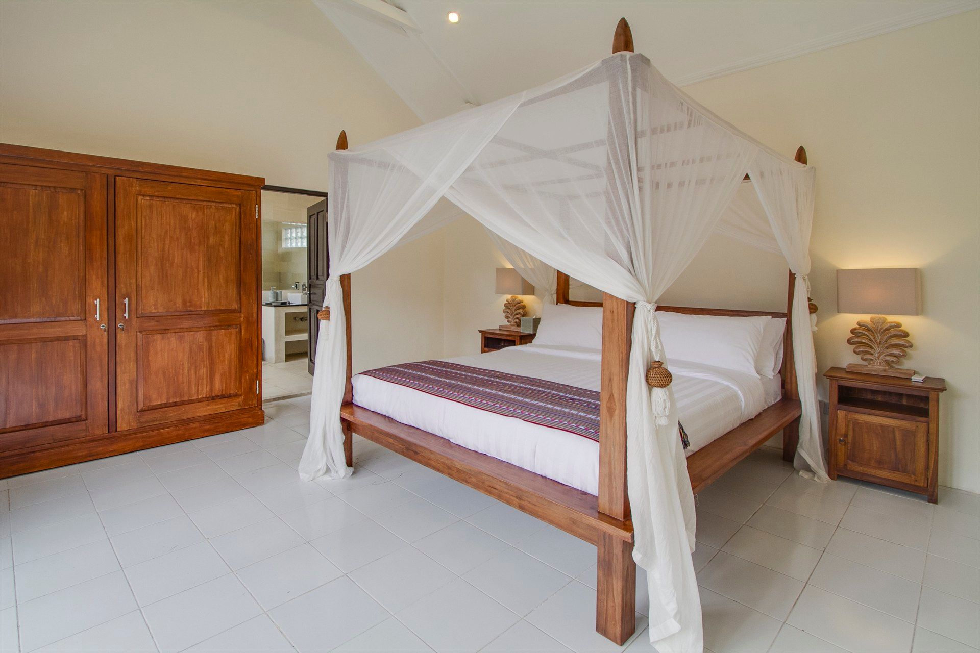 Bedroom with four-poster bed, white bedding, wooden furniture, and light-colored walls and floor.