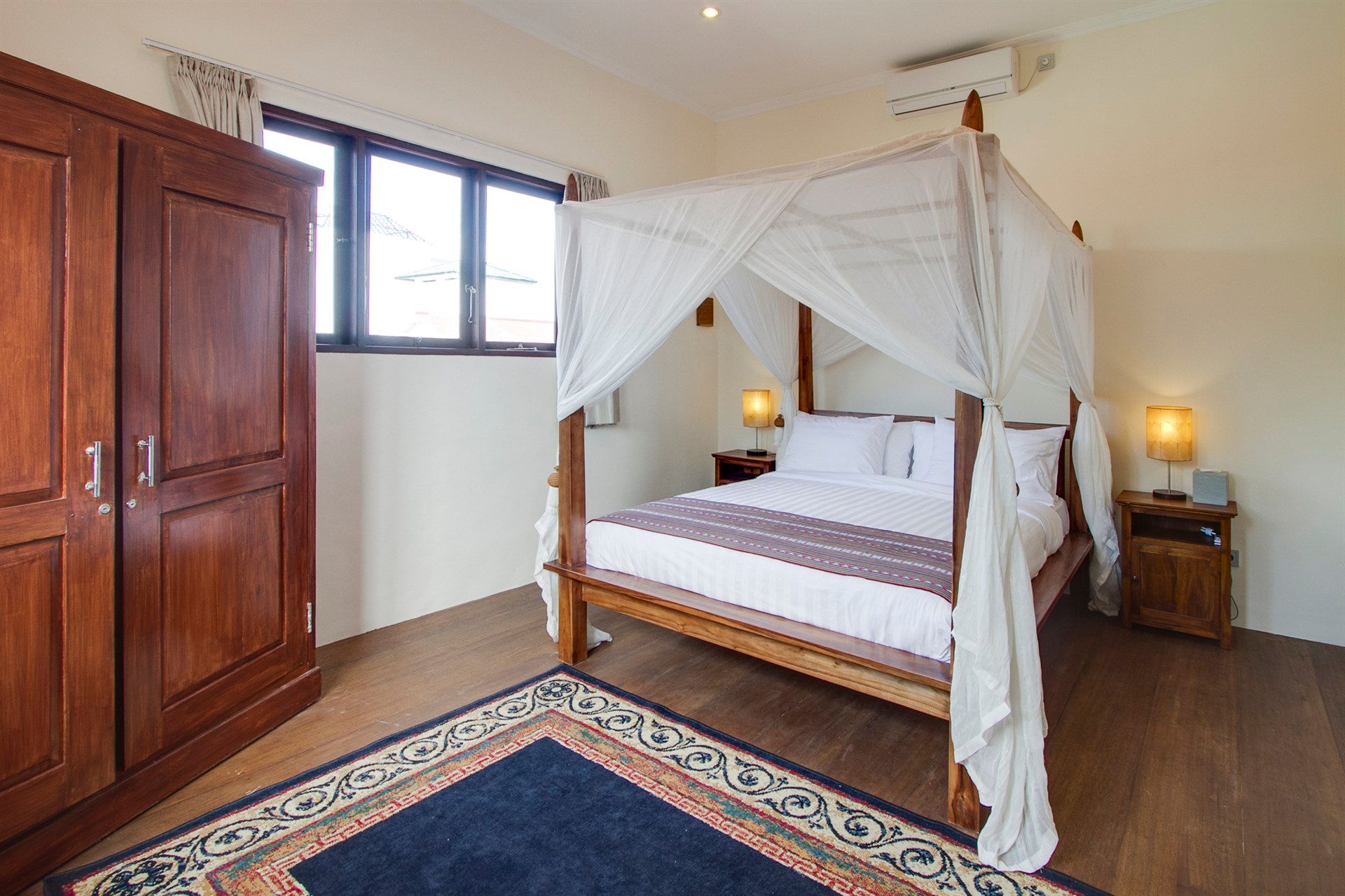 Bedroom with canopy bed, wooden furniture, blue rug, and window.