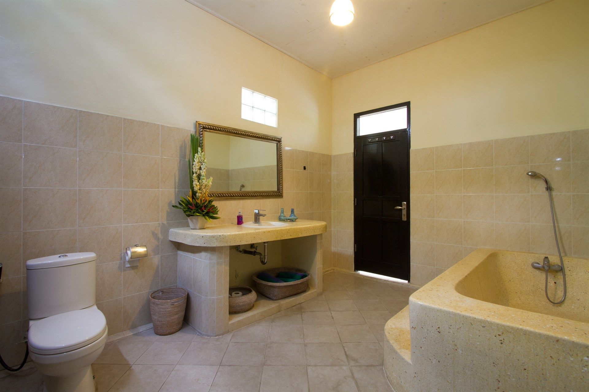 Bathroom with stone-like tile and fixtures: toilet, vanity, and bathtub. Beige walls and a dark door.