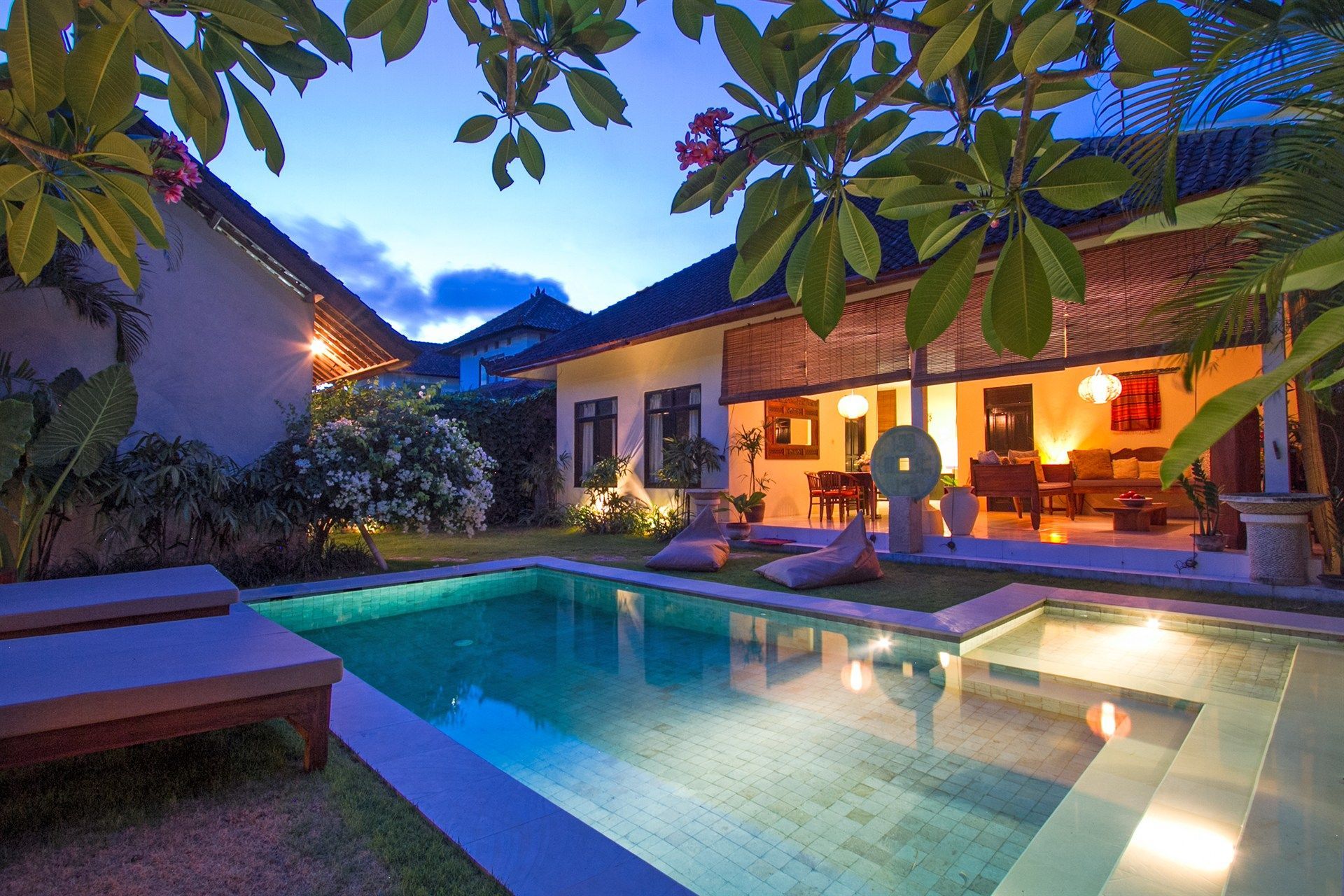 Night view of a villa with a pool, lush greenery, and illuminated living room.
