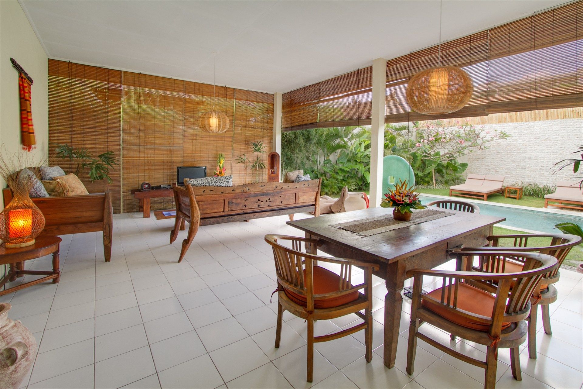 Open-air dining space with wooden furniture, overlooking a pool. Bamboo blinds, and a neutral color palette.